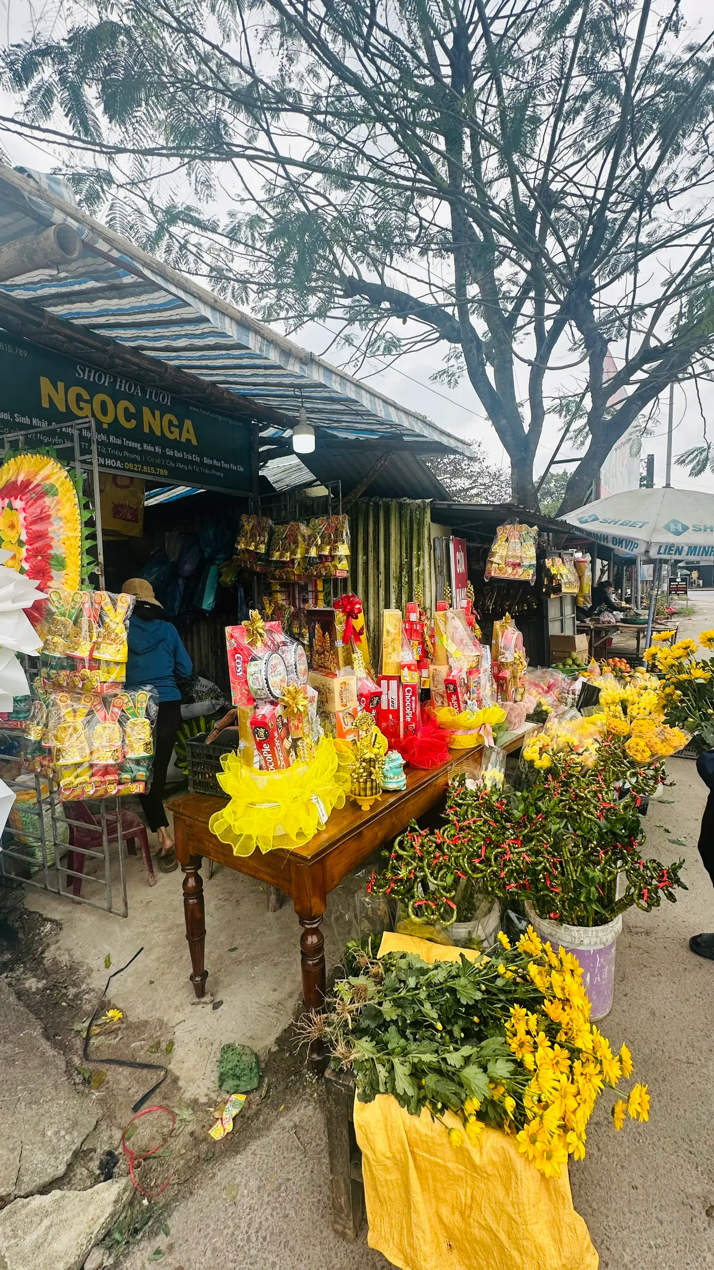 Góc hoa tươi và đồ lễ trước tiệm Ngọc Nga, cúc vàng và lá xanh
