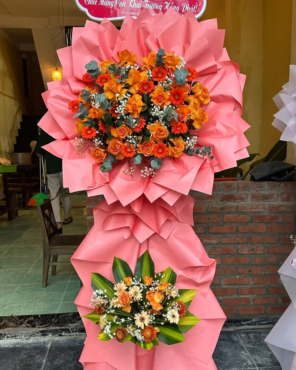 Kệ hoa khai trương hai tầng tông cam, hoa hồng cam và cúc trên giấy gói hồng